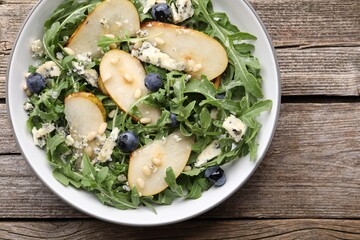 Tasty salad with arugula, pear, blue cheese, peanuts and blueberries in bowl on wooden table, top view