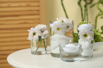 Burning candles and flowers in glasses of water on white table indoors