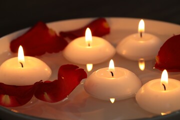 Burning candles and petals in bowl of water on table, closeup
