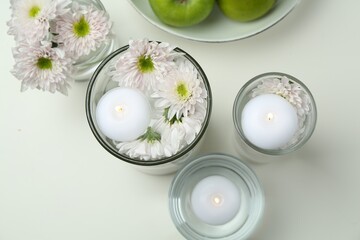 Burning candles and flowers in glasses of water on white table, flat lay
