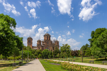 Tasmajdan Park in Belgrade with St. Mark Orthodox Church in neo-Byzantine style, surrounded by green trees, flower beds and benches under summer sky.

