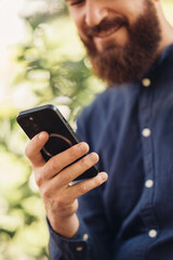 A smiling bearded man happily engages with his smartphone while surrounded by lush green foliage and nature