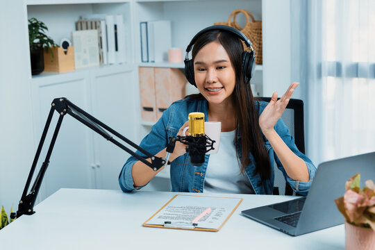 Host channel Asian influencer talking in broadcast streaming online wearing headset on social media live with script note, greeting listeners in life coaching or marketing at modern studio. Stratagem.