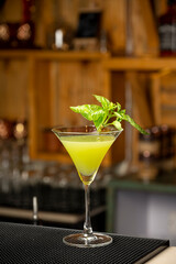 Close-up shot of green cocktail served in a martini glass, garnished with fresh mint, with blurred bar background
