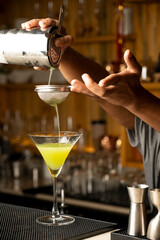 Close-up shot of a bartender’s hand pouring a green cocktail through a sieve into a martini glass, with a blurred bar background

