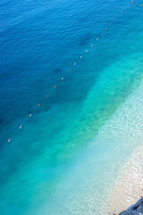 Crystal clear turquoise sea from above. Aerial view of crystal clear turquoise sea water gently meeting a sandy shore, with a floating buoy line dividing swimming areas.

