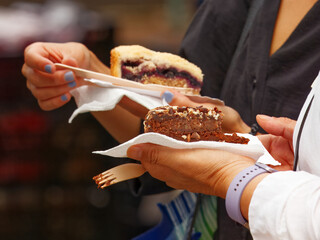 Two women holding slices of fresh cakes, enjoying sweet desserts in a relaxed weekend market atmosphere.