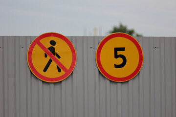 A "No Pedestrians" Sign And A "5" Speed Limit Sign, Both Yellow And Red, On A Corrugated Metal Fence At A Construction Or Industrial Site.