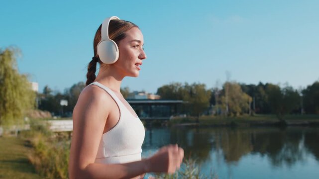 Side view of athletic fitness sport runner girl training, putting on wireless headphones and listening favorite music song. Workout cardio in morning city park. Young woman jogging healthy lifestyle.