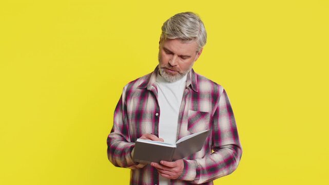 Thoughtful journalist Caucasian middle-aged man making notes, writing down thoughts with pen into notepad notebook diary, to do list, good idea. Mature old guy isolated on yellow background indoors