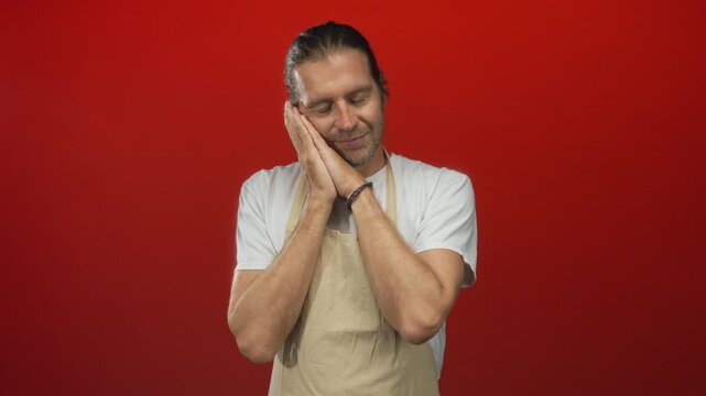 Man with long hair wearing a beige apron folds hands in prayer posture in a red studio setting; serenity.