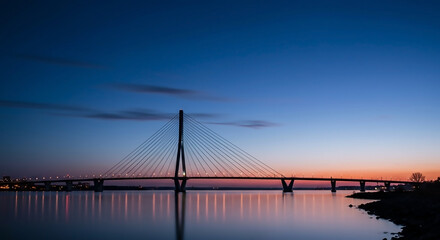 A modern cable-stayed bridge illuminated at dusk, with its structure and city lights beautifully reflected in the serene water.