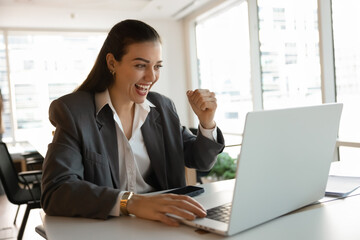 Business success. Overjoyed excited young businesswoman entrepreneur project manager office worker raise hand up scream yes great look at notebook screen celebrate startup growth remote work victory