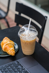 Take away coffee in a plastic cup and fresh croissant, working at cafe