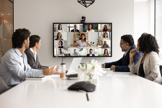 Modern tech innovation for business communication. Diverse multiracial team collaborate during hybrid online and in person meeting for briefing event interact both at conference table and by videocall