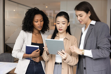 Informal idea sharing and casual teamwork. Three beautiful young women coworkers from diverse ethnic groups meet in office stand together talk engaged in creative brainstorming collaborate with tablet