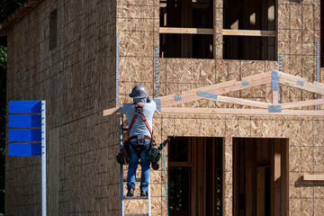 Carpenter on ladder with hard hat and tool belt using a hammer to move a wood frame rafter joist into place, new house construction project
