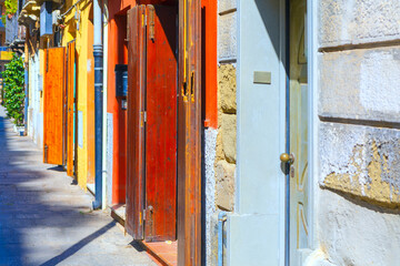 Row of colorful building doors with open wooden shutters lines narrow European street. Bright sunlight creates deep shadows on cobblestone pavement. Folding doors stand out against toned walls