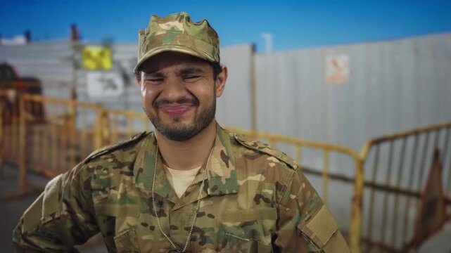 Man in camouflage uniform grimaces at construction fencing near building; duty pressure frustration.