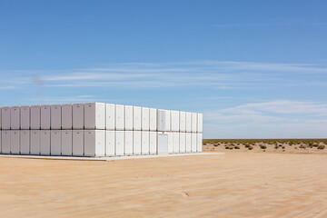 A large stack of white building blocks or insulation panels sits in a sandy desert landscape under a clear blue sky with wispy clouds