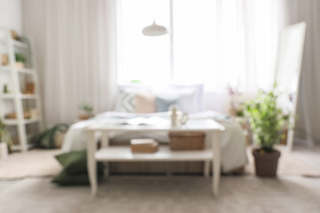 Blurred view of light bedroom with tables, shelf unit and plants