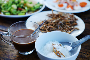 A glass cup of hot coffee with crispy fried Anchovy fish intestines on white dish and boiled rice. Thai food on wooden table is breakfast in the morning of Thailand
