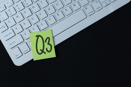 An overhead shot of a white computer keyboard and a sticky note with text "Q3". Business quarters, financial planning, and quarterly reports.