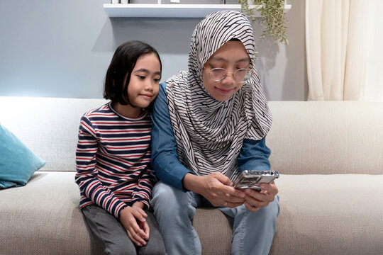 A mother wearing a hijab and her daughter looking at the phone. Modern family moment of sharing and technology use in a comfortable, home setting.
