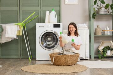 Young woman with wicker basket and bottles of detergent sitting near modern washing machine in...