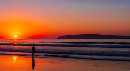 A lone person watches a vibrant orange sunset over the ocean from the wet, sandy shore.