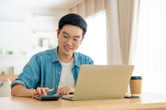 Happy young Asian man working on laptop in living room. Freelance worker male sitting at desk writing notes while watching webinar, studying online
