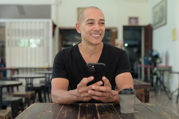 Confident Bald Hispanic man in black t-shirt sitting in outdoors cafe restaurant using phone
