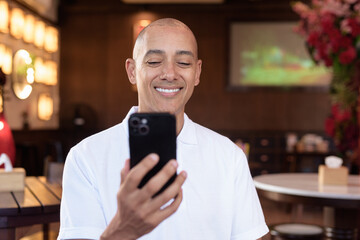 Confident Bald Hispanic man in white polo shirt sitting in Chinese style coffee shop using phone