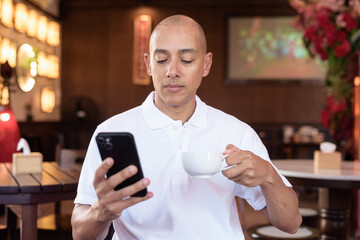 Confident Bald Hispanic man in white polo shirt sitting in Chinese style coffee shop using phone