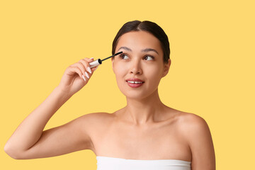 Fototapeta premium Beautiful young Asian woman applying mascara on yellow background