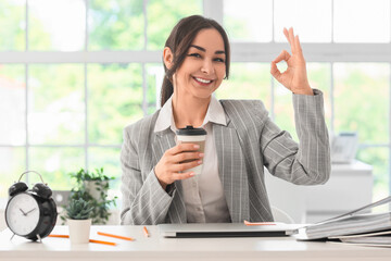 Businesswoman with coffee showing OK at table in office. Time management concept