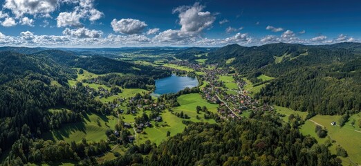 Panoramic view of a valley with a lake and villages nestled in the mountains. Lush green fields and forests surround a tranquil alpine scene under a partly cloudy sky