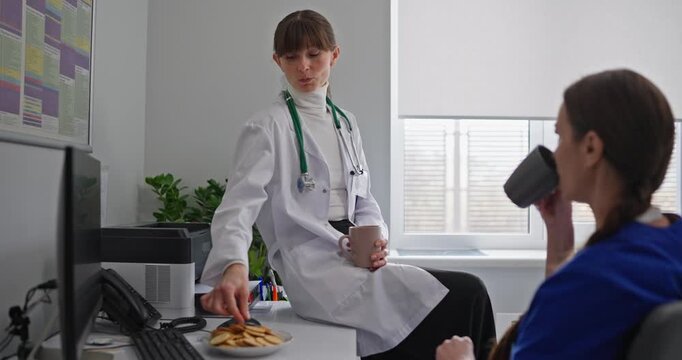 Female doctor chatting with her colleague and eating snacks during lunch in hospital office. Two female doctors chat and drink tea during their break from hospital work