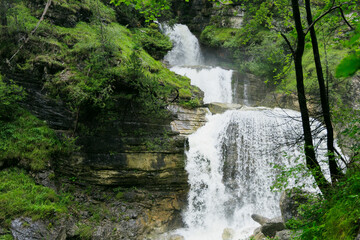 Stunning view of the multi-tiered Kuhflucht Gorge waterfall in Farchant, Bavaria, Germany. The cascading water flows down steep rock formations surrounded by lush green forest, showcasing the natural 