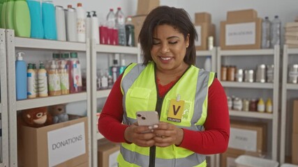 Woman volunteer wearing a reflective vest uses her phone in a charity donation center room with organized shelves of supplies.