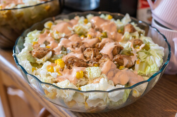 Large glass bowl of grilled chicken salad with lettuce, corn, pickles, and creamy pink dressing, served on a wooden surface in a home kitchen.