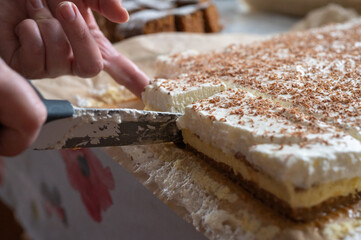 Close-up of a hand cutting a homemade cake topped with whipped cream and grated chocolate, served on parchment paper.
