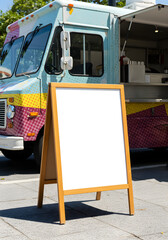 A blank wooden A-frame sign stands on the pavement in front of a colorful food truck.