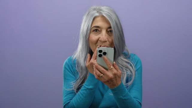 Senior woman with grey hair using smartphone against purple background, expressing surprise and amusement in isolated vibrant setting.