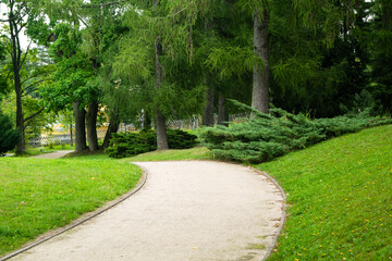 A peaceful, winding gravel path curves through a lush green park, flanked by manicured bushes and mature trees. This image evokes feelings of a relaxing walk, nature, and tranquility.