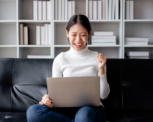 Young Adult Woman Cheering with Laptop Success on Sofa in Living Room  Achieving Goals Freelancer or Online Learning at Home with Bookshelf Background