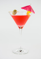 A red cocktail in a classic martini glass with a green olive, an umbrella and a twist of orange peel set against a white background.