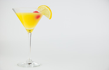 An orange cocktail in a classic martinin glass with a wedge of lemon and red cocktail cherry set against a white background.