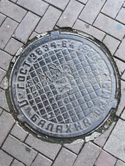 Top Down View Of An Old Cast Iron Manhole Cover With Soviet GOST Standard Marking On A Paving Stone Sidewalk. Vertical Urban Detail.