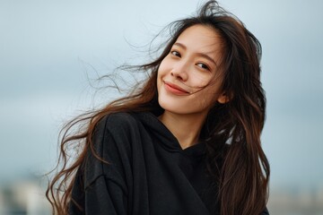 Fototapeta premium Young woman with long hair enjoying a breezy day outdoors while wearing a hoodie against a cloudy sky backdrop, smiling happily in an urban setting